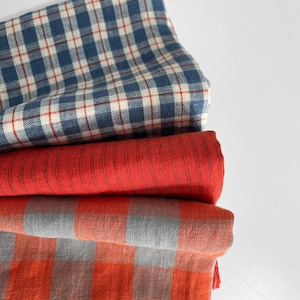 May include: A collection of folded fabrics in different patterns and colours. The top layer features a blue and white check with red detailing. Beneath it is a solid red fabric, followed by a grey and orange check, and another orange and grey check.
