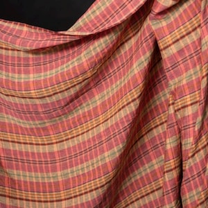 May include: A draped fabric with a plaid pattern in shades of pink, red, brown, and green. The fabric is folded and flowing, with visible horizontal and vertical lines creating the plaid design. The background is black.