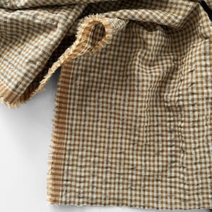 May include: A piece of fabric featuring a brown and white gingham check pattern. The textile has a crinkled, textured surface and unfinished, frayed edges along the sides, showcasing the weave of the material in a folded, layered arrangement.