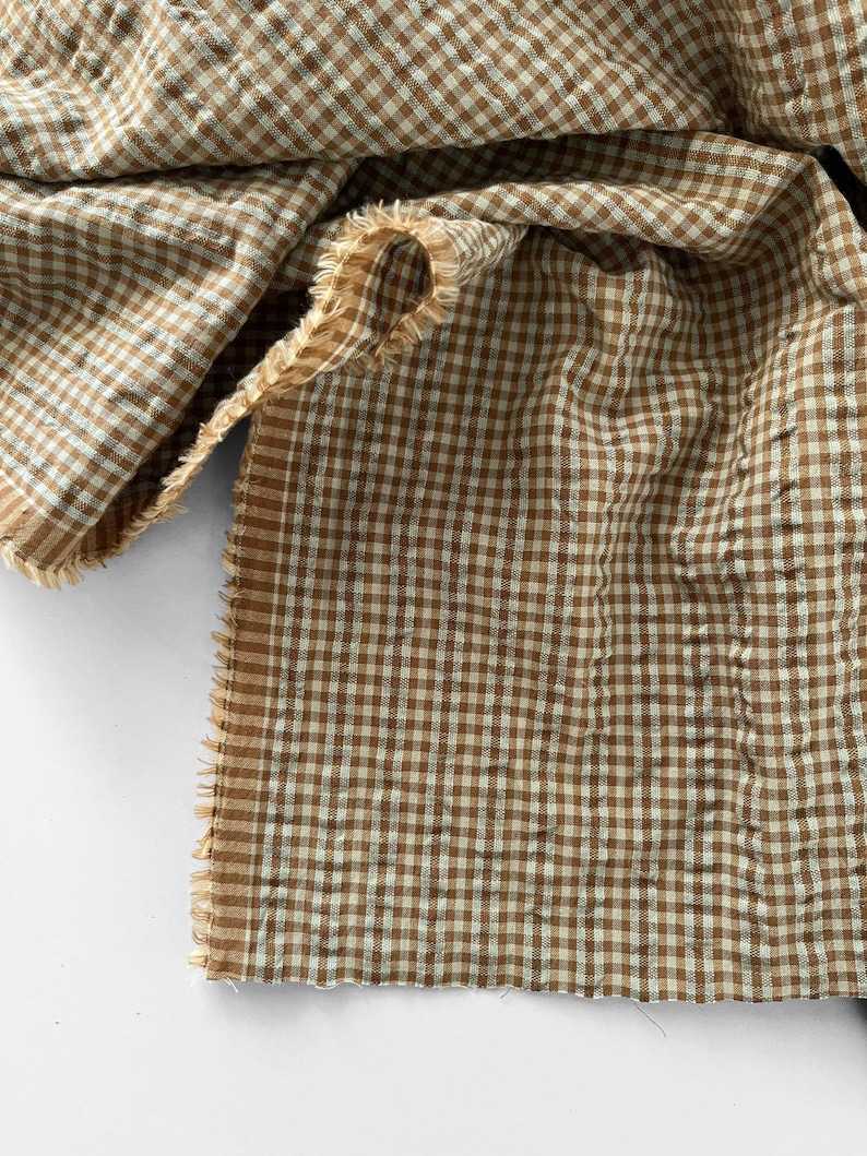 May include: A piece of fabric featuring a brown and white gingham check pattern. The textile has a crinkled, textured surface and unfinished, frayed edges along the sides, showcasing the weave of the material in a folded, layered arrangement.