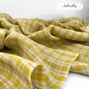May include: A close-up of a yellow plaid fabric with a pink grid pattern. The fabric is draped and folded, creating soft shadows. The word "daffodilly" is written in a white oval.