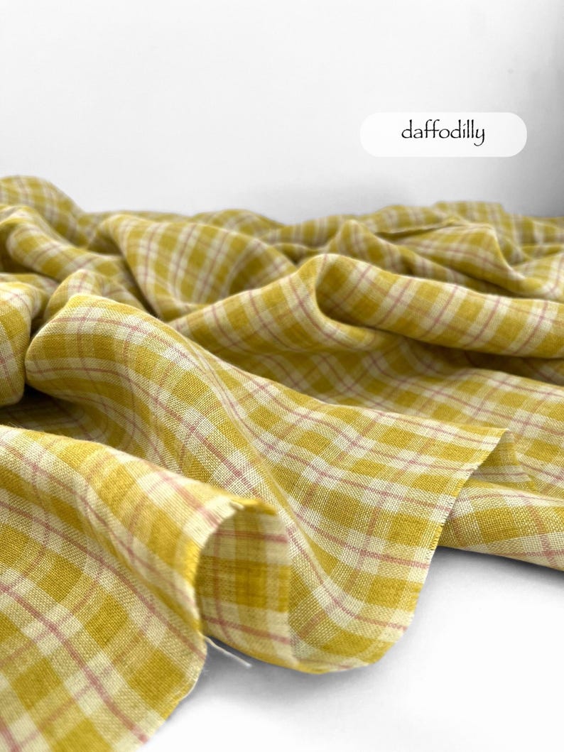 May include: A close-up of a yellow plaid fabric with a pink grid pattern. The fabric is draped and folded, creating soft shadows. The word "daffodilly" is written in a white oval.