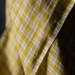 May include: Close-up of a folded fabric with a yellow and white plaid pattern, accented by thin pink lines. The fabric has a soft, textured appearance, with visible folds and shadows that add depth to the image. The overall aesthetic is rustic and inviting.
