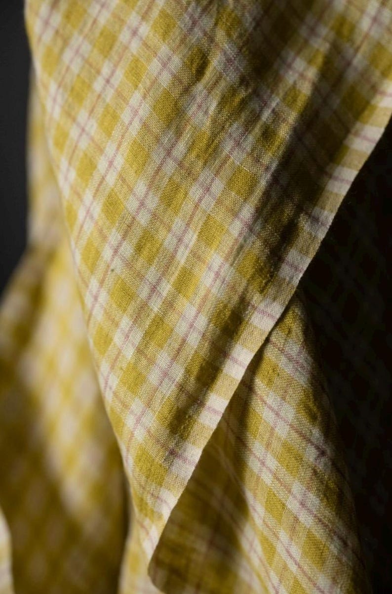 May include: Close-up of a folded fabric with a yellow and white plaid pattern, accented by thin pink lines. The fabric has a soft, textured appearance, with visible folds and shadows that add depth to the image. The overall aesthetic is rustic and inviting.