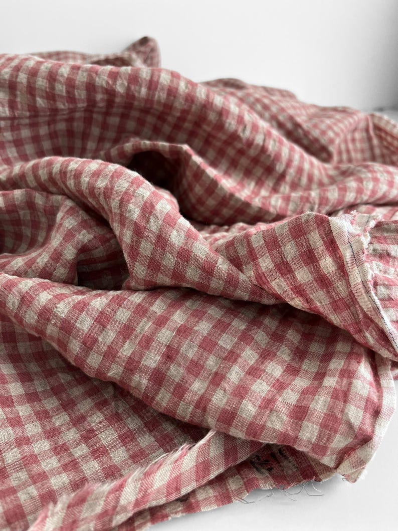 May include: A close-up of a red and white gingham fabric. The fabric is draped and folded, creating a textured appearance. The pattern is a classic checkered design, with the red and white squares evenly spaced.
