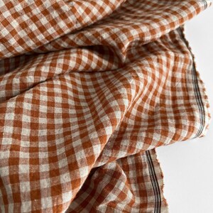 May include: Close-up of a folded fabric with a classic gingham pattern in shades of rust and cream. The fabric has a soft, textured appearance, with visible weave details. The edges are slightly frayed, adding to its rustic charm.