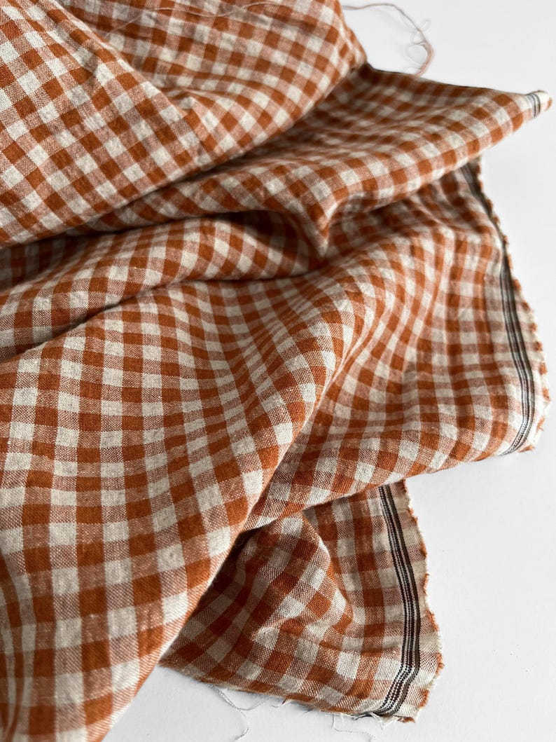 May include: Close-up of a folded fabric with a classic gingham pattern in shades of rust and cream. The fabric has a soft, textured appearance, with visible weave details. The edges are slightly frayed, adding to its rustic charm.
