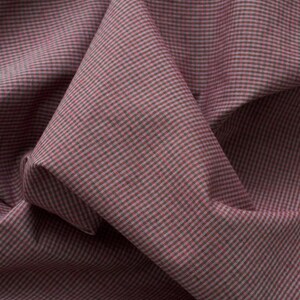 May include: A piece of fabric featuring a fine, repeating micro-checkered pattern in muted shades of dusty rose pink and dark charcoal grey. The textile is draped to show soft folds and creases, highlighting the consistent grid-like weave texture.