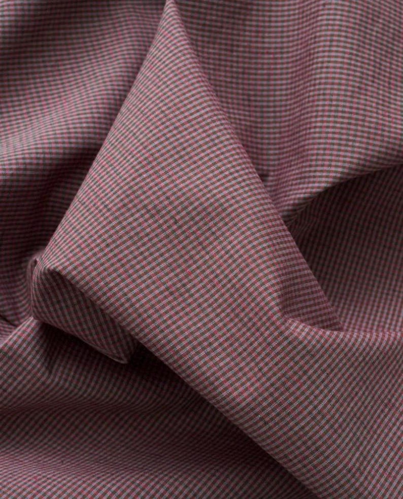 May include: A piece of fabric featuring a fine, repeating micro-checkered pattern in muted shades of dusty rose pink and dark charcoal grey. The textile is draped to show soft folds and creases, highlighting the consistent grid-like weave texture.