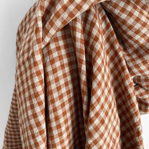 May include: Close-up of a draped fabric with a classic gingham pattern. The fabric features a repeating square design in a warm orange-brown and off-white color scheme. The material appears soft and flowing, with visible folds and texture.