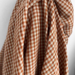May include: Close-up of a fabric with a classic gingham pattern. The fabric features a repeating pattern of small squares in a warm, earthy orange and off-white color scheme. The fabric is draped, showing the texture and folds.