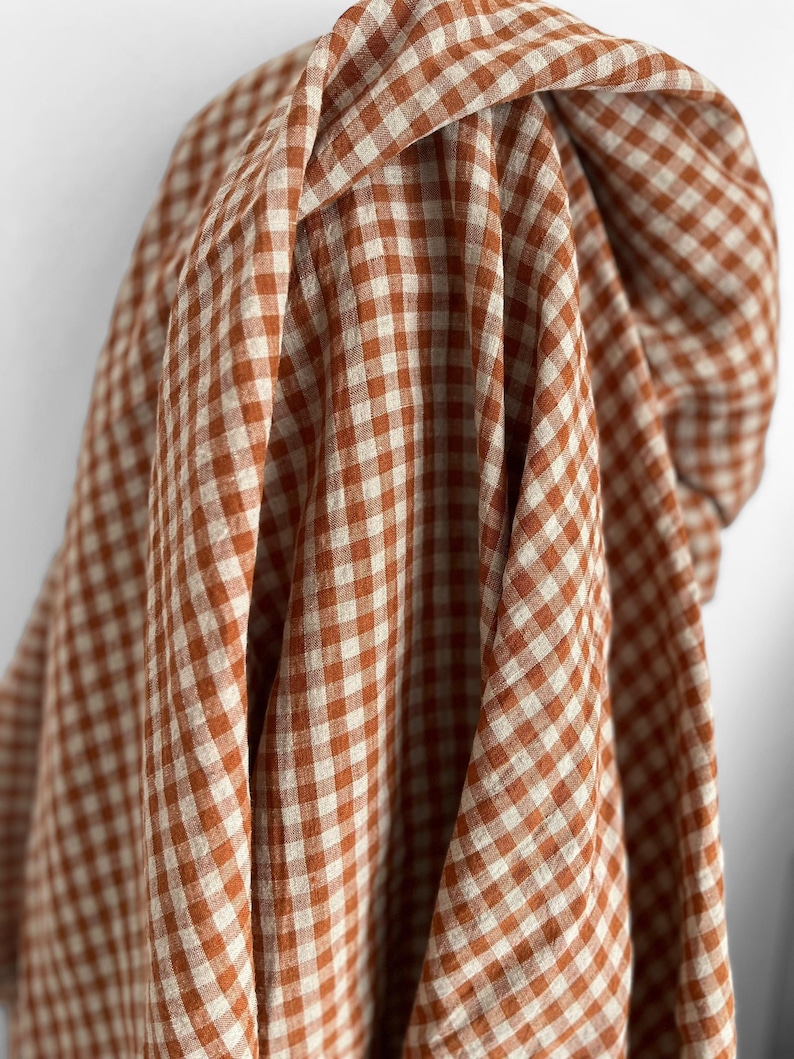 May include: Close-up of a fabric with a classic gingham pattern. The fabric features a repeating pattern of small squares in a warm, earthy orange and off-white color scheme. The fabric is draped, showing the texture and folds.