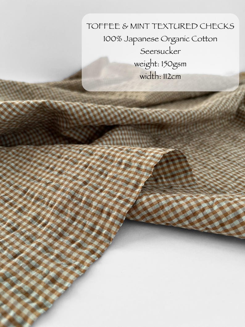May include: Toffee and mint coloured seersucker fabric featuring a small checkered pattern. The textile is labelled as 100% Japanese organic cotton with a weight of 150gsm and a width of 112cm. The material has a puckered, textured surface.