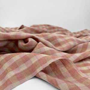Puede incluir: Primer plano de una tela doblada con un patrón de cuadros rosa y beige. La tela está drapeada, mostrando su textura y tejido. La paleta de colores es suave y apagada, con una sensación natural y orgánica.