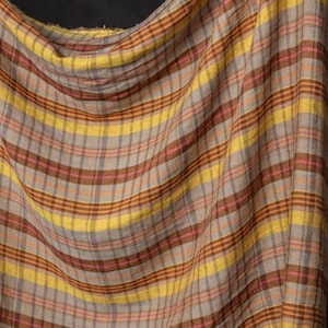 May include: A draped fabric with a plaid pattern. The design features alternating stripes of yellow, brown, pink, and gray. The fabric is soft and flowing, with a textured appearance.