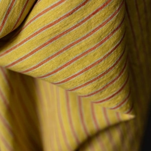 May include: Close-up of a folded fabric with a yellow background and thin, parallel red stripes. The fabric has a textured appearance, with the stripes running vertically and horizontally across the folds. The overall effect is a warm, inviting textile.