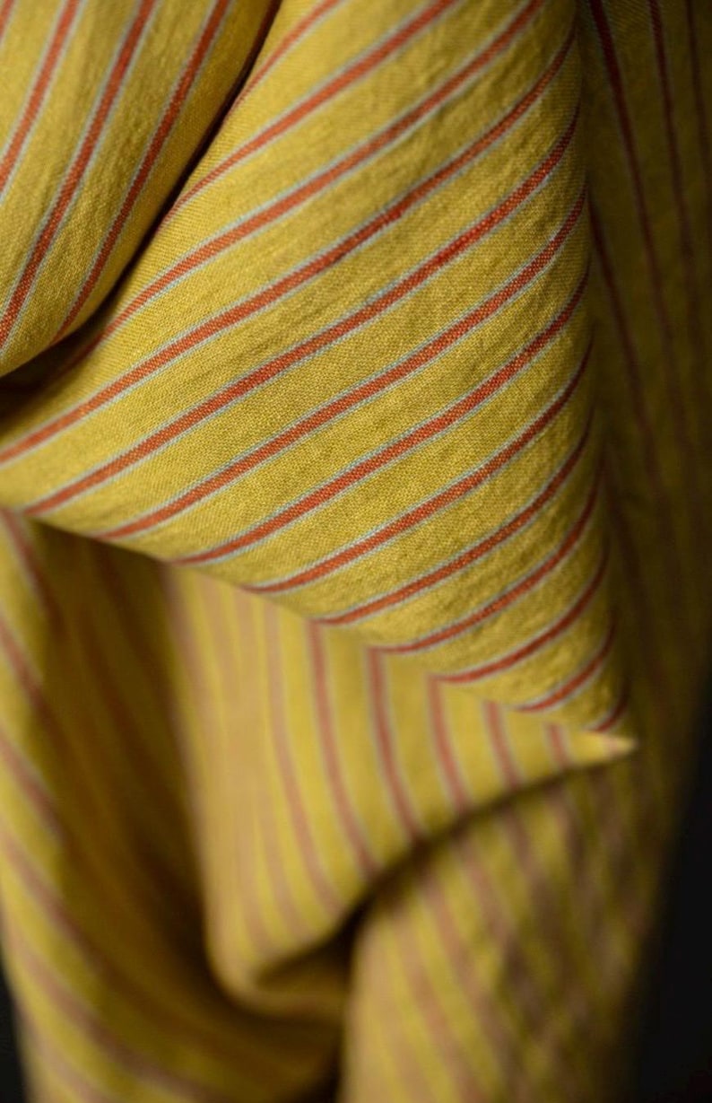 May include: Close-up of a folded fabric with a yellow background and thin, parallel red stripes. The fabric has a textured appearance, with the stripes running vertically and horizontally across the folds. The overall effect is a warm, inviting textile.