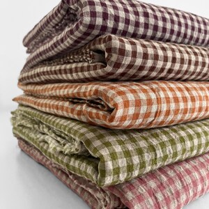 May include: A stack of folded fabric in various colors, including burgundy, brown, orange, green, and pink. Each piece of fabric features a classic gingham pattern, creating a rustic and inviting aesthetic. The fabrics appear to be linen or a similar natural material.