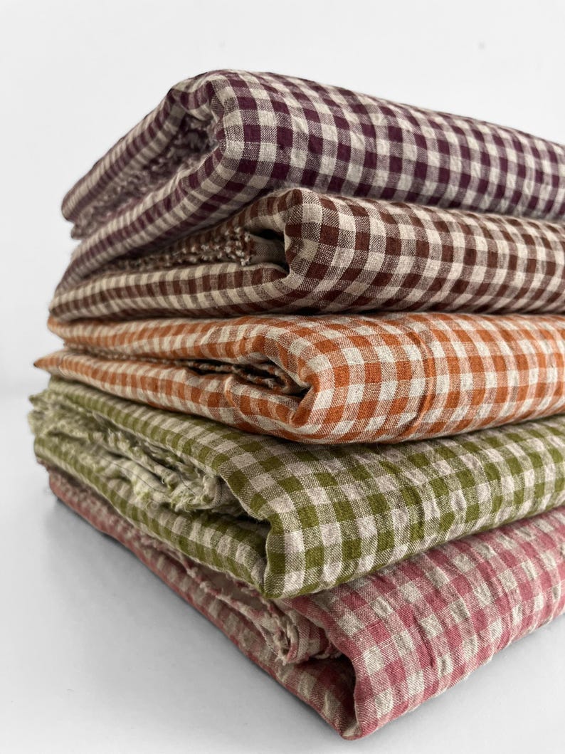 May include: A stack of folded fabric in various colors, including burgundy, brown, orange, green, and pink. Each piece of fabric features a classic gingham pattern, creating a rustic and inviting aesthetic. The fabrics appear to be linen or a similar natural material.