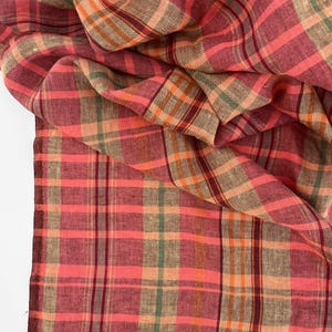 May include: A close-up of a plaid fabric in shades of red, pink, orange, green, and brown. The fabric is folded and draped, showcasing the pattern's intricate lines and color variations. The material appears to be lightweight and possibly linen.
