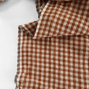 May include: Close-up of a fabric with a classic gingham pattern in shades of brown and cream. The fabric is folded, revealing the texture and weave. The edges are unfinished, showing the raw cut of the material. Ideal for home decor or apparel.