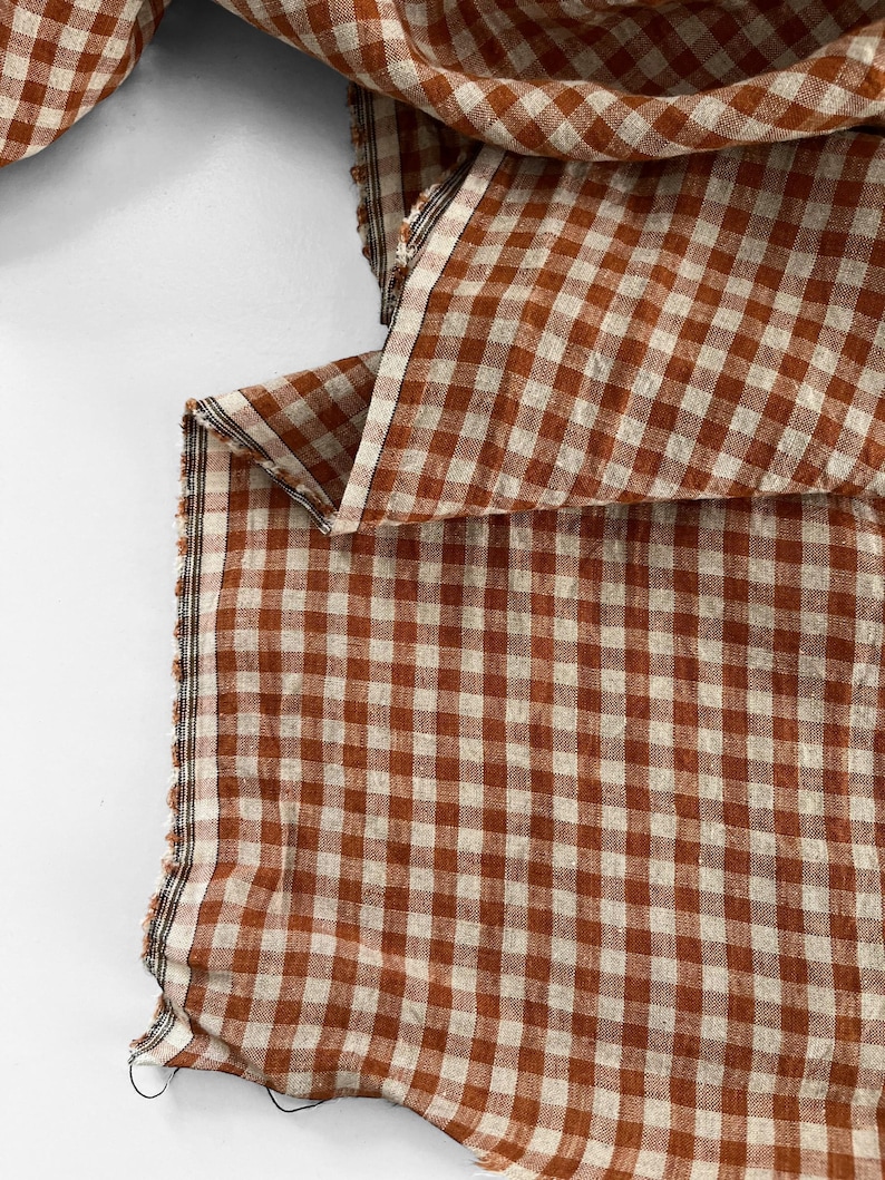 May include: Close-up of a fabric with a classic gingham pattern in shades of brown and cream. The fabric is folded, revealing the texture and weave. The edges are unfinished, showing the raw cut of the material. Ideal for home decor or apparel.