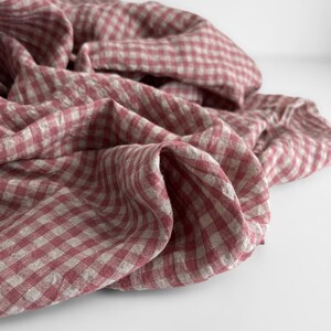 May include: A close-up shot of a folded fabric with a red and white gingham pattern. The fabric is draped, creating soft folds and shadows. The material appears to be linen or a similar lightweight textile, suitable for home decor or apparel.
