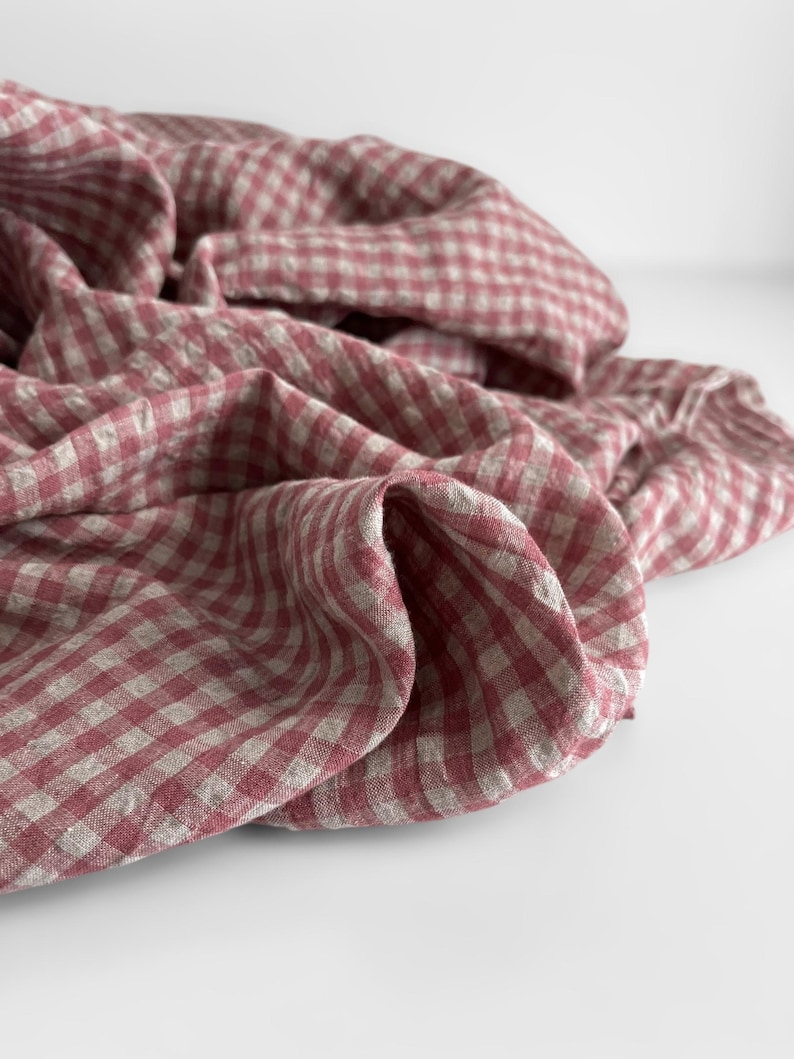 May include: A close-up shot of a folded fabric with a red and white gingham pattern. The fabric is draped, creating soft folds and shadows. The material appears to be linen or a similar lightweight textile, suitable for home decor or apparel.