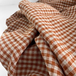 May include: Close-up of a folded fabric with a classic gingham pattern. The fabric features a warm, burnt orange and white checkered design. The cloth is draped, showing the texture and folds of the material. Ideal for home decor or crafting projects.