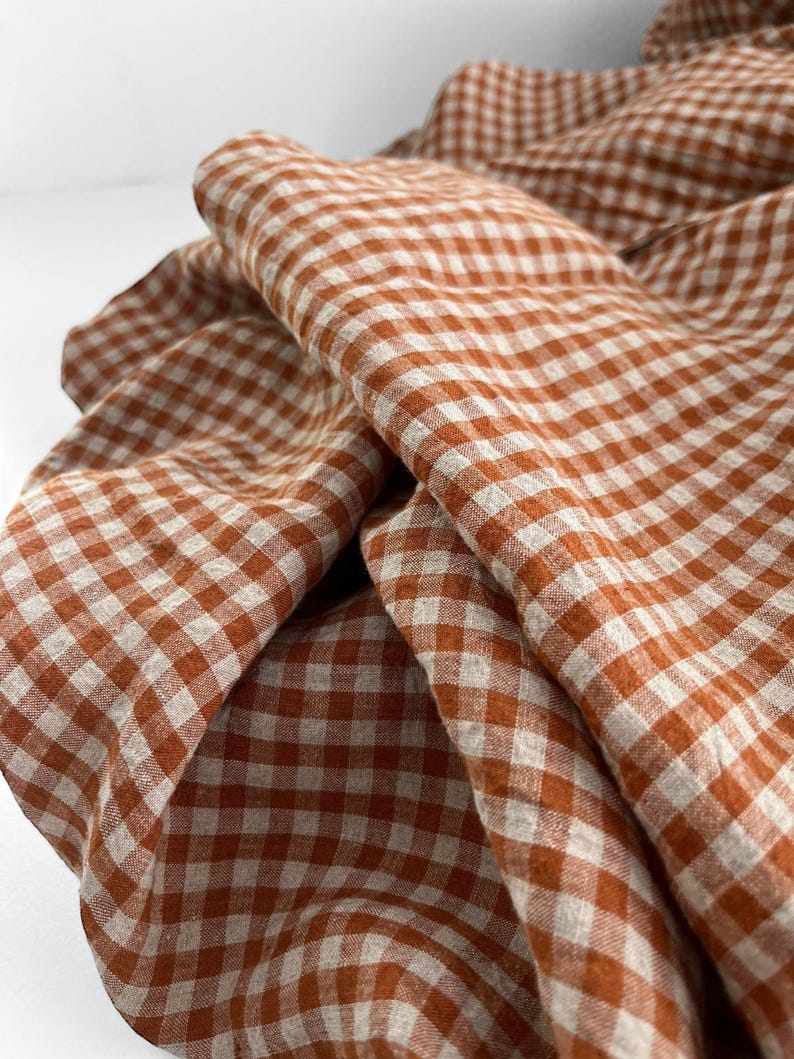 May include: Close-up of a folded fabric with a classic gingham pattern. The fabric features a warm, burnt orange and white checkered design. The cloth is draped, showing the texture and folds of the material. Ideal for home decor or crafting projects.