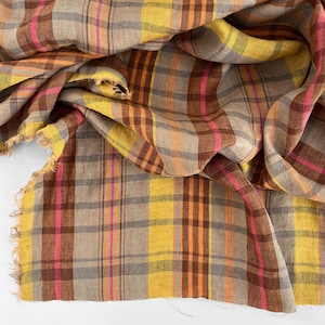 May include: A close-up of a plaid fabric in shades of brown, yellow, pink, and orange. The fabric has a fringed edge and is draped in soft folds. The pattern is a classic plaid design, suitable for apparel or home decor.