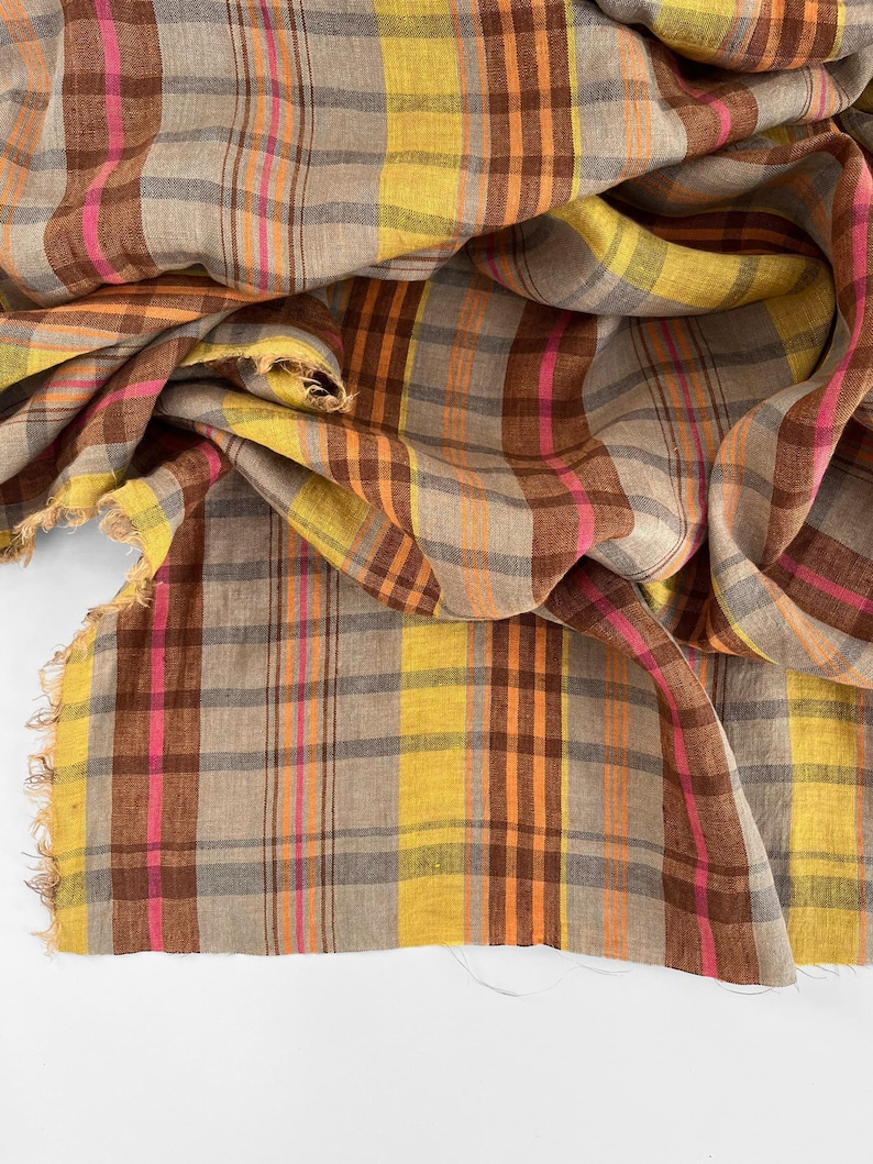 May include: A close-up of a plaid fabric in shades of brown, yellow, pink, and orange. The fabric has a fringed edge and is draped in soft folds. The pattern is a classic plaid design, suitable for apparel or home decor.