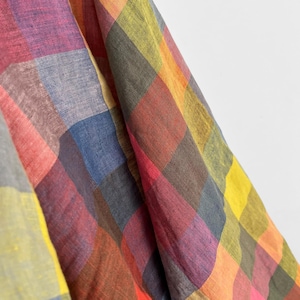 May include: Close-up of a colorful plaid fabric. The fabric features a large-scale plaid pattern in a variety of colors, including red, pink, purple, blue, yellow, and orange. The fabric appears to be soft and flowing.