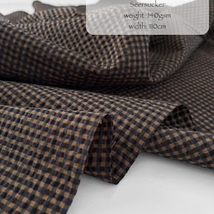 May include: A brown and black mini check seersucker fabric made of 100% Japanese organic cotton. The textile features a puckered texture and is labelled as WOODS MINI CHECKS, 100% Japanese Organic Cotton Seersucker, weight: 140gsm, width: 110cm.