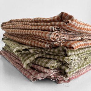 May include: A stack of folded fabric cloths in shades of orange, green, and pink. Each cloth features a checkered pattern. The edges of the cloths appear to be frayed, giving them a rustic appearance.