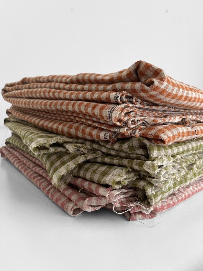 May include: A stack of folded fabric cloths in shades of orange, green, and pink. Each cloth features a checkered pattern. The edges of the cloths appear to be frayed, giving them a rustic appearance.