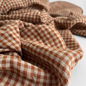 May include: Close-up of a folded fabric with a gingham pattern in shades of rust and cream. The fabric is draped, showing the texture and weave of the material. The pattern is a classic, small-scale check.