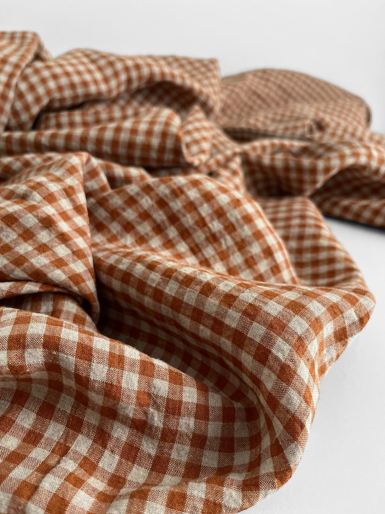 May include: Close-up of a folded fabric with a gingham pattern in shades of rust and cream. The fabric is draped, showing the texture and weave of the material. The pattern is a classic, small-scale check.