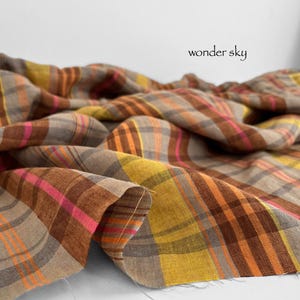 May include: Close-up of a fabric with a plaid pattern in shades of brown, orange, yellow, and pink. The fabric is draped, with the text "wonder sky" in black script.
