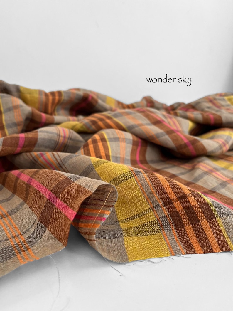 May include: Close-up of a fabric with a plaid pattern in shades of brown, orange, yellow, and pink. The fabric is draped, with the text "wonder sky" in black script.