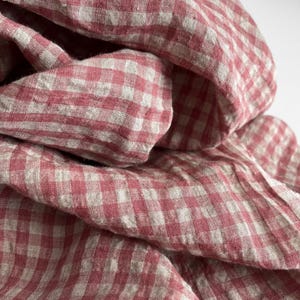 May include: Close-up of a folded fabric with a red and white gingham pattern. The fabric appears to be linen or a similar lightweight material, with a soft, textured appearance. The folds create shadows and depth.