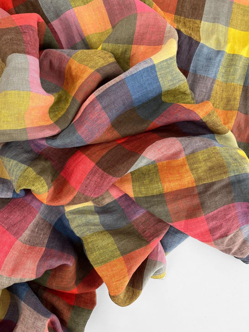 May include: A colorful, checkered fabric in shades of red, orange, yellow, green, blue, and brown. The fabric is draped and folded, showcasing the various colors and patterns. The material appears to be soft and lightweight, possibly linen or cotton.