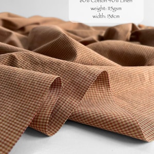 May include: Fabric with a sunset micro check pattern in shades of orange and cream. The text reads: SUNSET MICRO CHECKS, 60% Cotton 40% Linen, weight: 115gsm, width: 138cm. The material is draped in soft folds, showing a fine, woven texture.