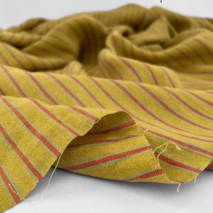 May include: Mustard yellow fabric with red and light blue stripes. The fabric is draped, showing the texture and pattern. The word "patisserie" is printed in black on the upper right corner.