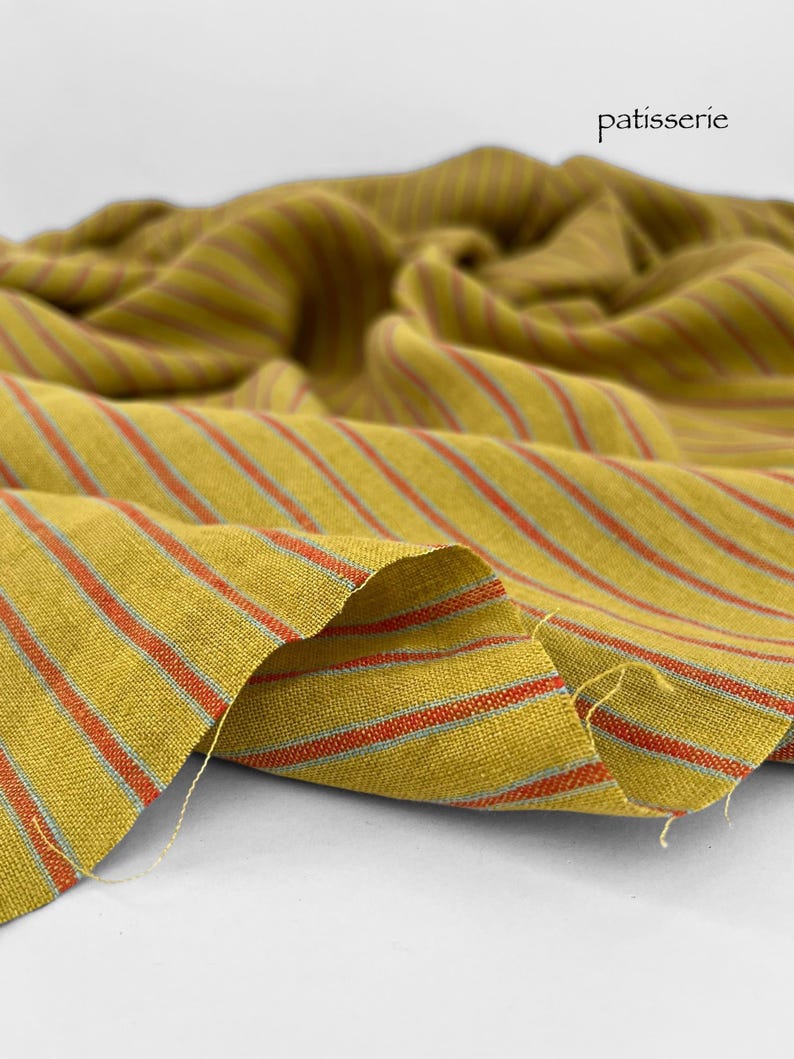 May include: Mustard yellow fabric with red and light blue stripes. The fabric is draped, showing the texture and pattern. The word "patisserie" is printed in black on the upper right corner.