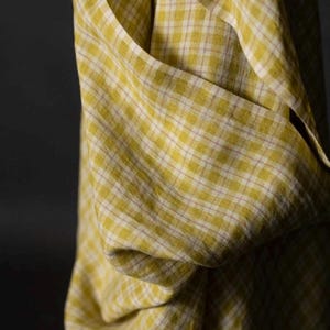 May include: Close-up of a yellow and white plaid fabric. The fabric is draped, showing folds and texture. The pattern is a classic check design, with a soft, muted color palette. The fabric appears to be lightweight.