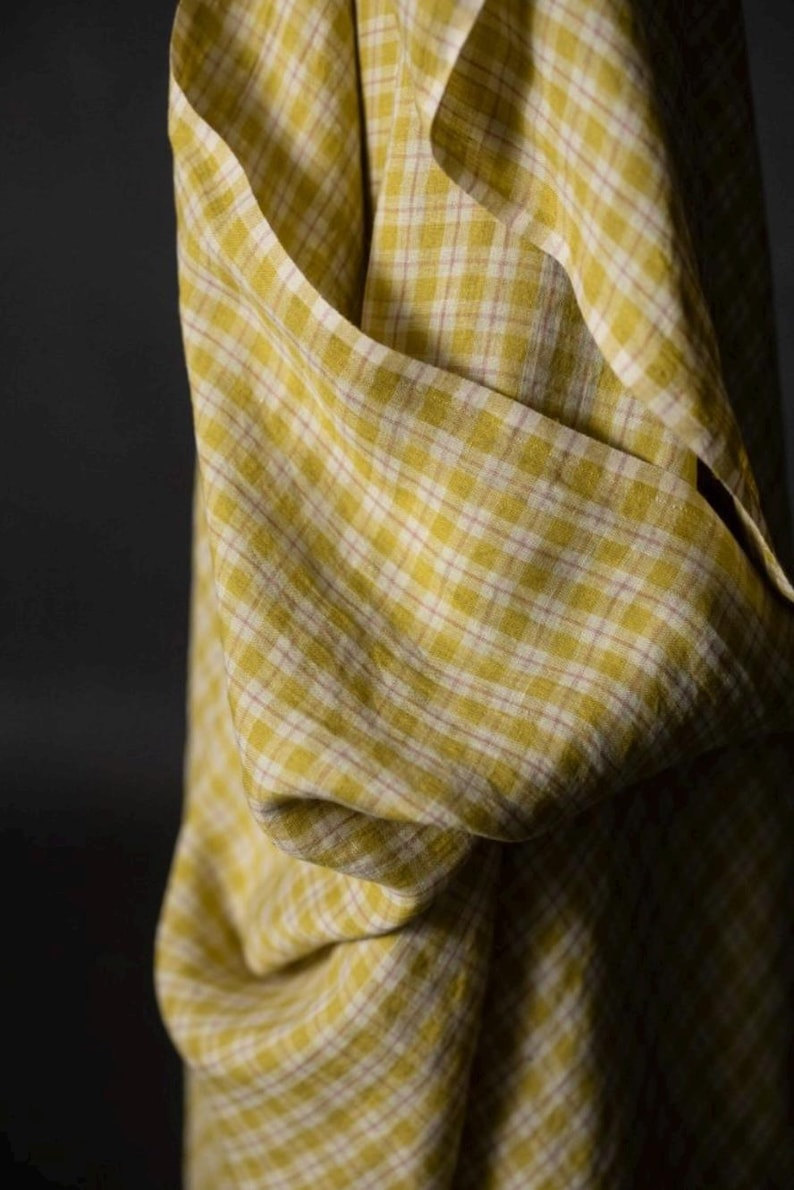 May include: Close-up of a yellow and white plaid fabric. The fabric is draped, showing folds and texture. The pattern is a classic check design, with a soft, muted color palette. The fabric appears to be lightweight.