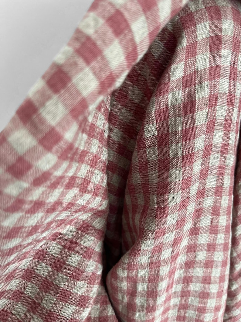May include: Close-up of a fabric with a pink and white gingham pattern. The fabric is folded, creating soft curves and shadows. The pattern is a classic, timeless design, perfect for apparel or home decor.