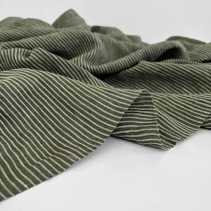 Op de afbeelding: Olijfgroene stof met dunne, witte horizontale strepen. De stof is gedrapeerd en gevouwen, waardoor zachte golven en schaduwen ontstaan. Het materiaal lijkt lichtgewicht en geschikt voor kleding- of woondecoratieprojecten. Afmetingen in centimeters.