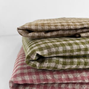 May include: A stack of three folded linen textiles in a gingham pattern. The top textile is a muted brown, the middle is olive green, and the bottom is a dusty rose color. The fabrics are neatly folded, showcasing the texture and pattern.